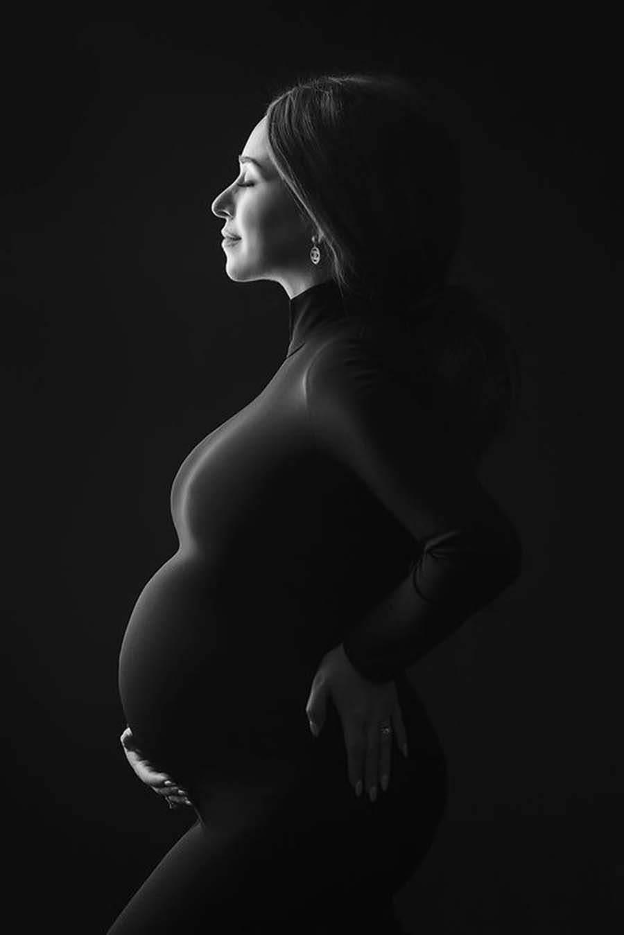Pregnant model poses on her side in a studio during a maternity photo session. She holds her back and bump. She wears a long and tight dress featuring turtle neck and long sleeves. Her eyes are closed and she looks away.