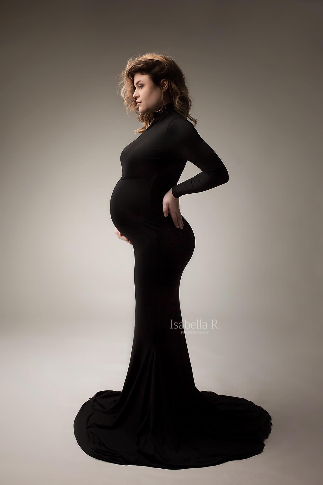 Short haired model poses on her side in a studio during a maternity photo session. She holds her back with one hand and her bump with the other. She wears a long and tight dress featuring turtle neck and long sleeves. Her eyes are open and she looks away. 