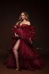 A woman is standing in a studio. She is wearing a big red tulle dress. The dress has a split at the front by her leg