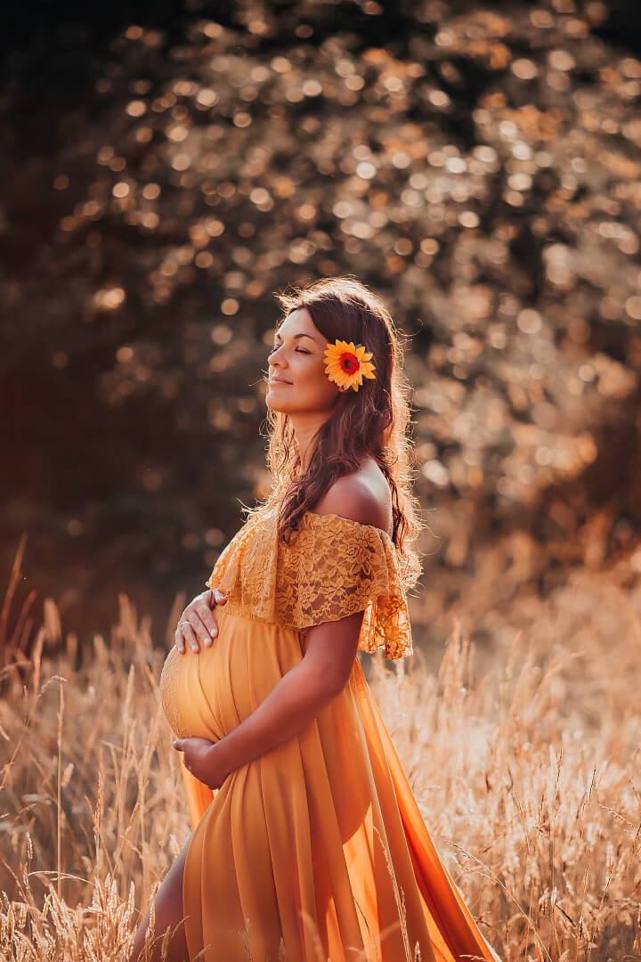 pregnant woman poses outside wearing a yellow maternity dress made of lace, jersey and chiffon. she has a sunflower on her head.