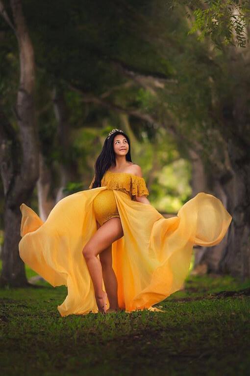 pregnant woman poses outside with a chiffon train dress in yellow color. she looks up and plays with the chiffon train.