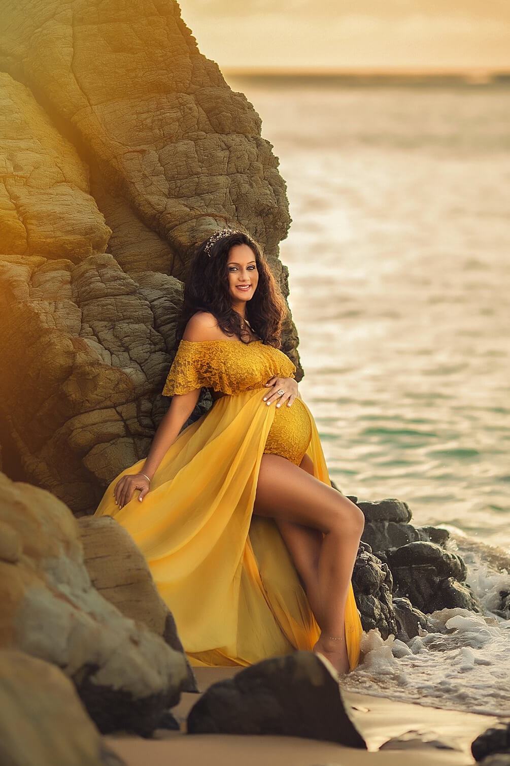 pregnant model poses by the water and smiling at the camera while wearing a long yellow dress and a crown.