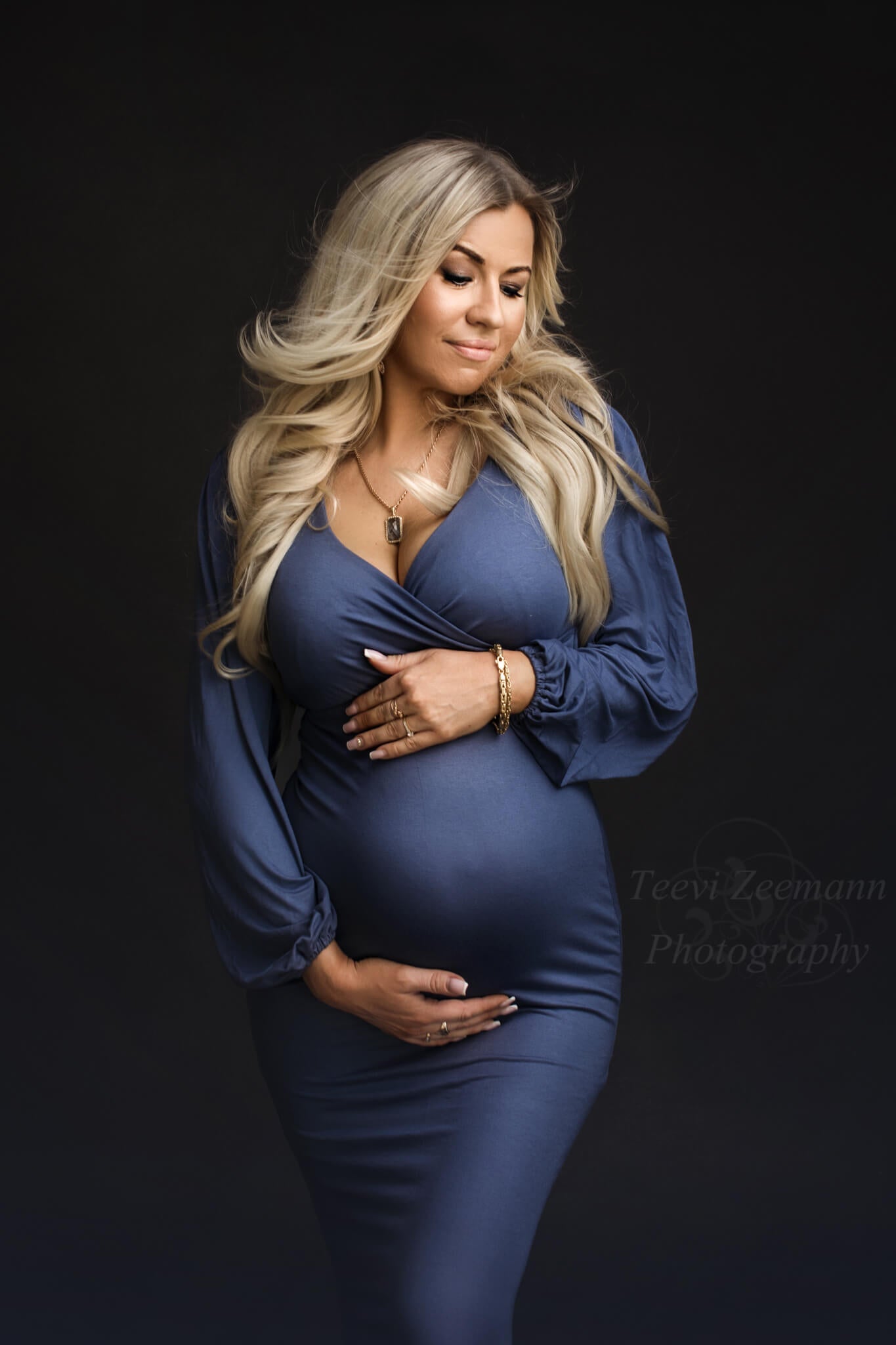 Blond pregnant model poses ina. studio wearing a denim dress made of jersey. She holds her bump with both hands and stares down. The dress has long bishop sleeves and an adjustable sweetheart neckline. 