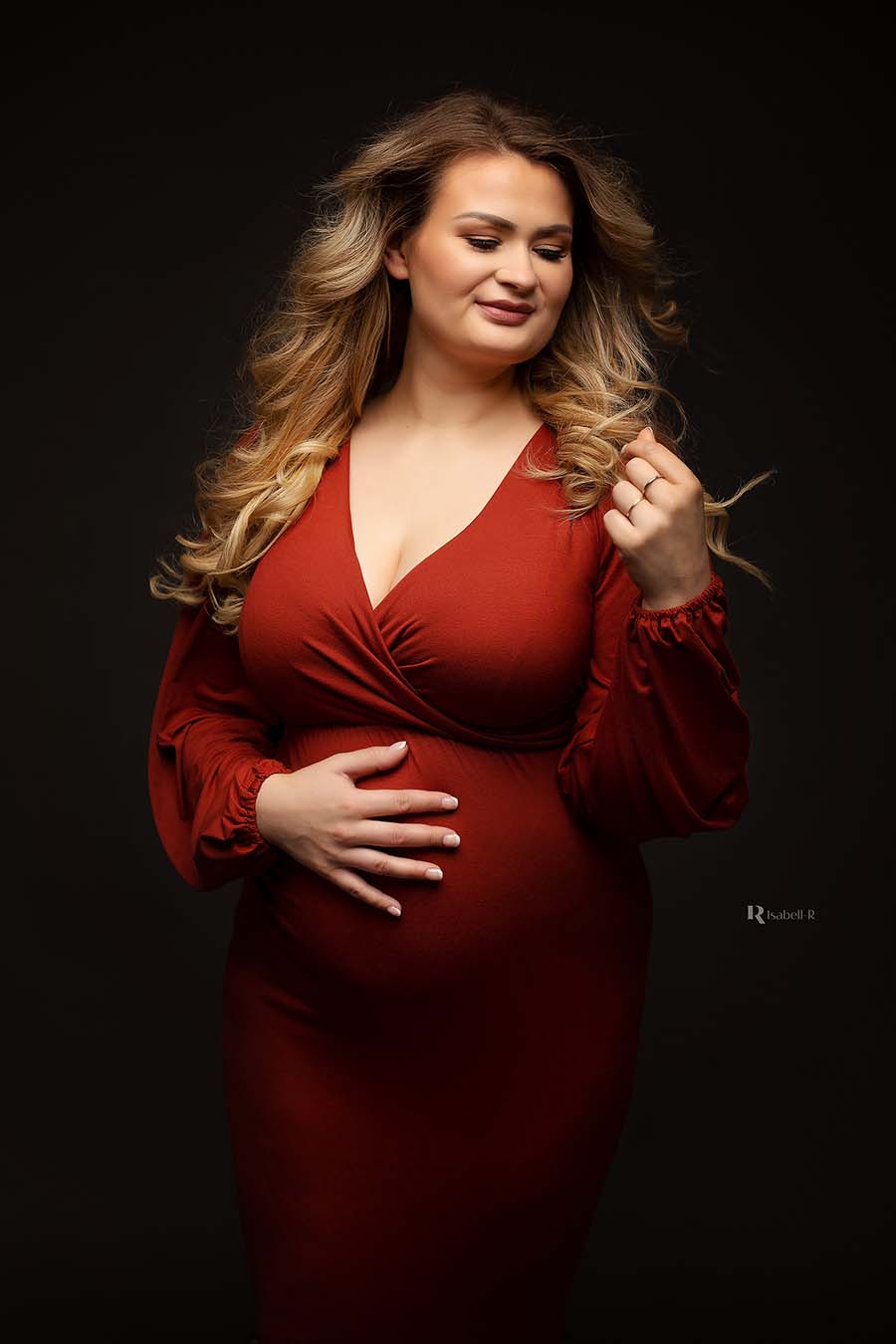 The dress the pregnant woman is wearing has a beautiful rust orange colour. She is looking at her hands. the dress has wide sleeves  and a sweetheart neckline