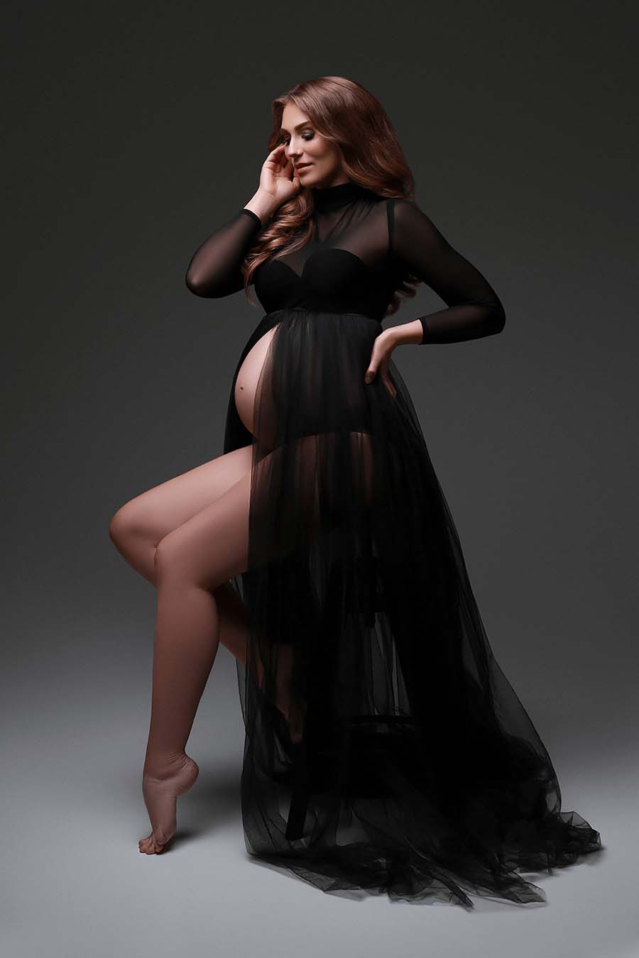 Red haired pregnant model poses in a studio wearing a tulle black dress with a turtleneck and long sleeves. The skirt is long and has a split in the middle. 