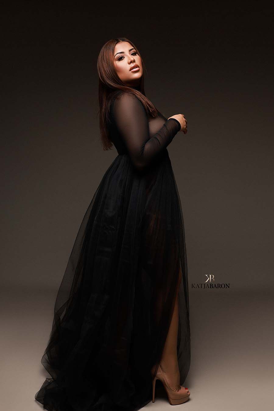 A pregnant woman is wearing a black dress and is see trough . The model is looking up. The dress has long sleeves and a big tulle skirt. 