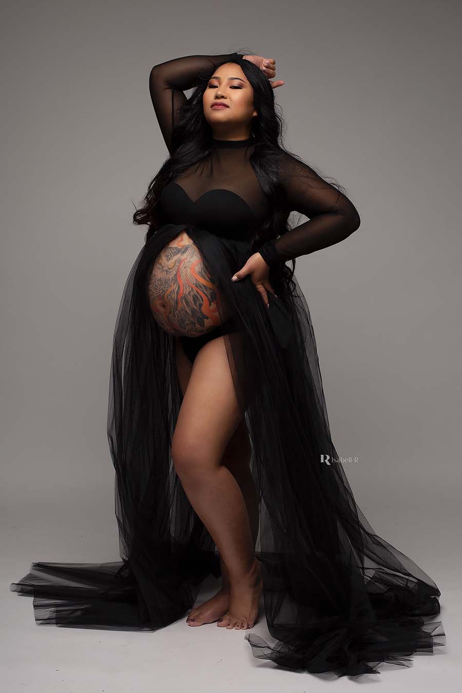 a woman is wearing a long black dress with a tulle skirt. The dress is open at the front . You can see her pregnant belly with a huge tattoo on it. She is wearing a black bra underneath