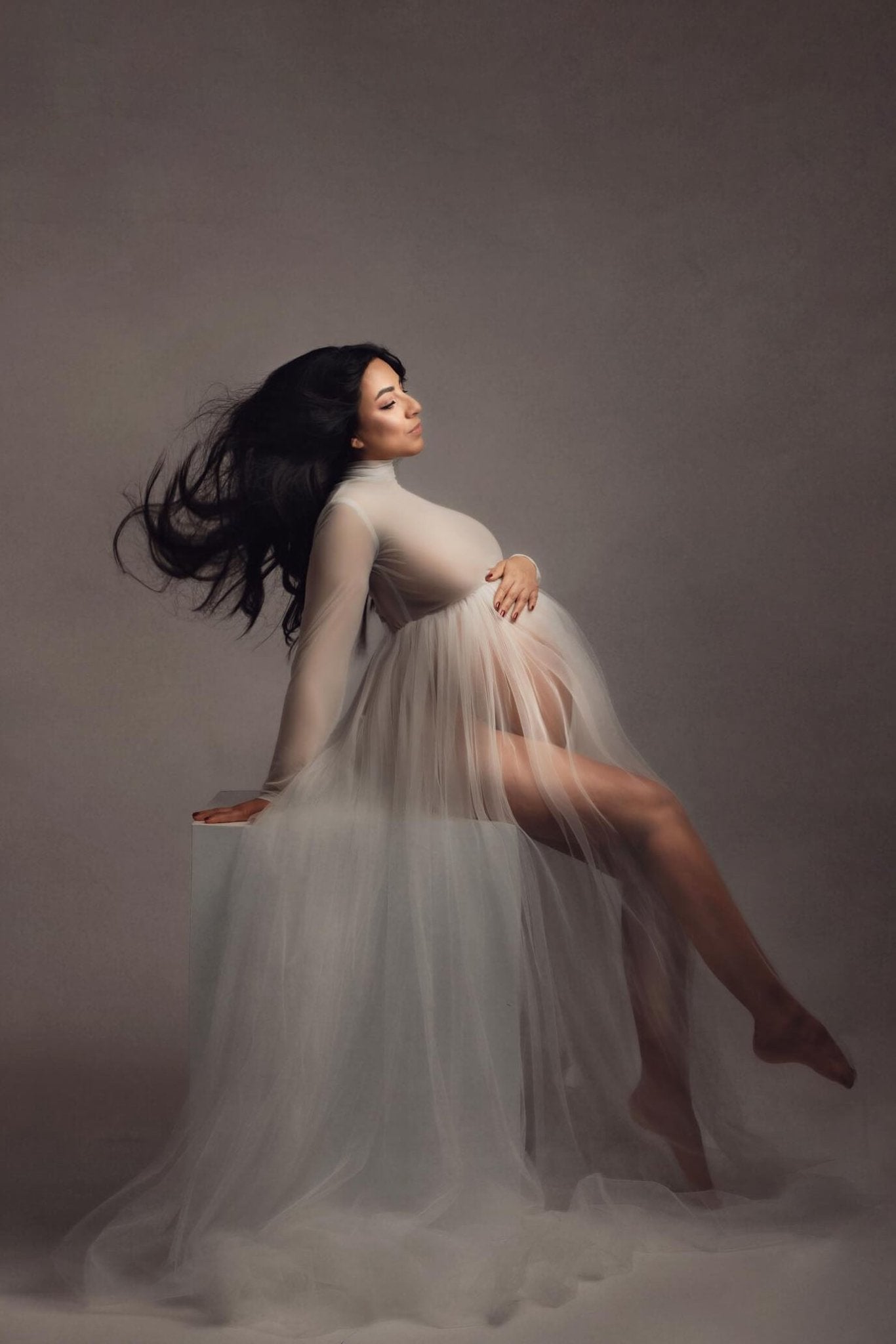 Long hair model poses in a studio during a maternity photoshoot. She sits while holds her bump with one hand. The photo is taken from her side and shows the tulle skirt and long sleeve turtleneck top she is wearing. Her outfit is off white. 