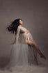 Long hair model poses in a studio during a maternity photoshoot. She sits while holds her bump with one hand. The photo is taken from her side and shows the tulle skirt and long sleeve turtleneck top she is wearing. Her outfit is off white. 