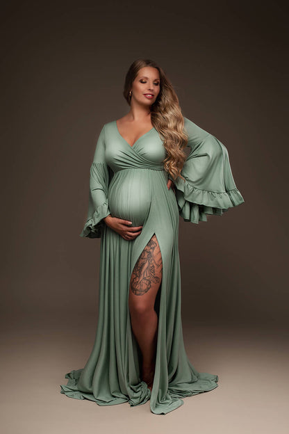 Blond pregnant model poses in a studio with an azur colored dress. The dress features long flared sleeves with ruffle details and an adjustable sweetheart neckline. The model holds her bump with one of the hands while the other lays on the side. She has her eyes partially closed and faces down to the camera. The dress can be completely seen. One of the models legs can be see through the side split in the skirt.
