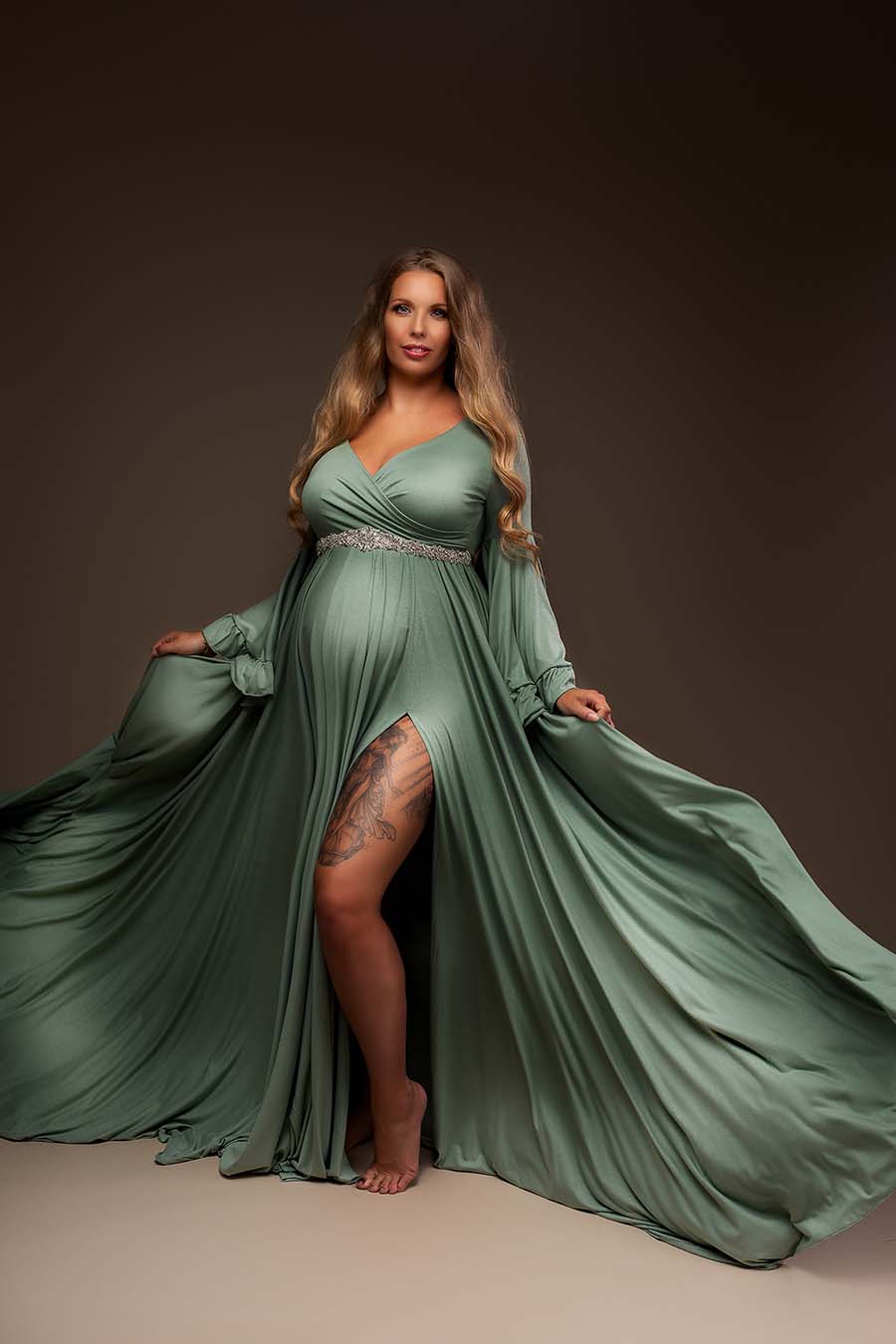 Blond pregnant model poses in a studio with an azur colored dress. The dress features long flared sleeves with ruffle details and an adjustable sweetheart neckline. The model holds with both hands the sides from the skirt so the wideness from the long skirt can be easily seen. She faces the camera and the dress can be almost completely seen. One of the models legs can be see through the side split in the skirt.