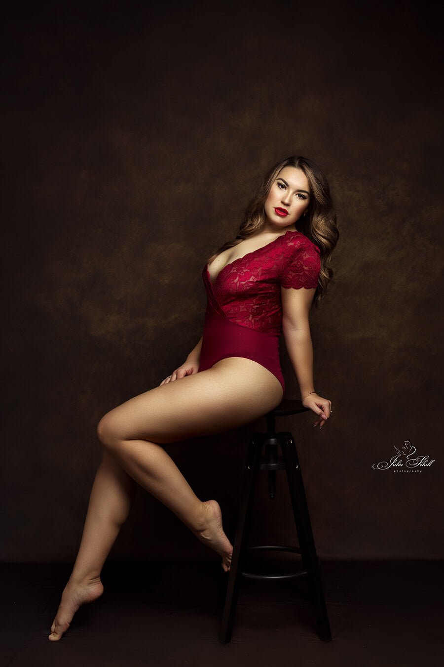 dark blond model poses in a studio, sitting on a high black chair. she is wearing a red bodysuit during a boudoir session.