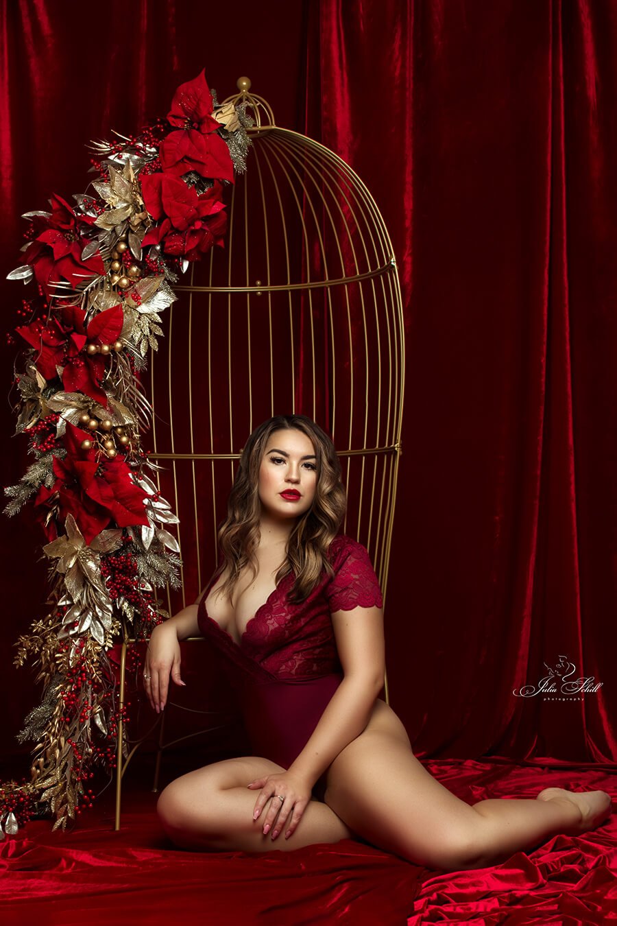 dark blond model poses in a studio, sitting next to a metal chair that resembles a cage with several red details.  she is wearing a red bodysuit during a boudoir session.