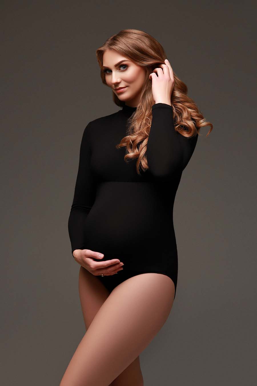 model stares to the camera while holding her hair and bump. she is wearing a black bodysuit with a turtle neck and long sleeves. 