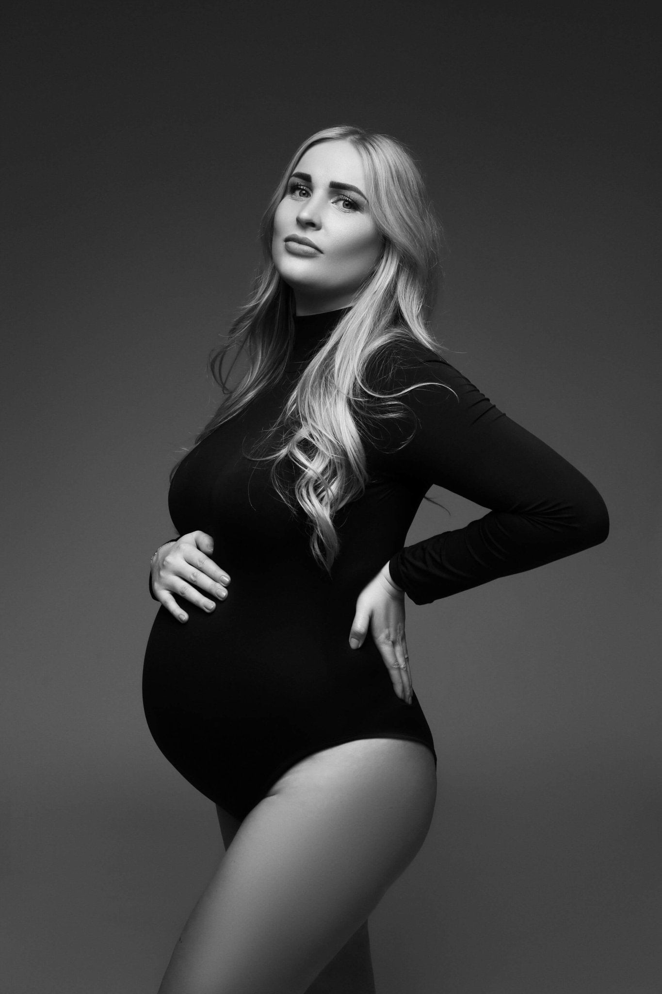 black and white close up in a studio during a maternity photoshoot. the model has long blond hair and is wearing a black bodysuit with a turtleneck and long sleeves. she is staring at the camera and holding her bump and waist. 