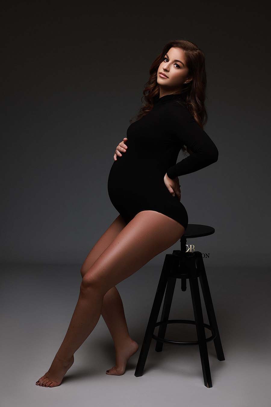 pregnant model is sitting on a stool during a maternity photoshoot in a studio. she is wearing a bodysuit with a turtleneck and long sleeves. she is looking to the camera while holding her bump and back. the bodysuit is black.