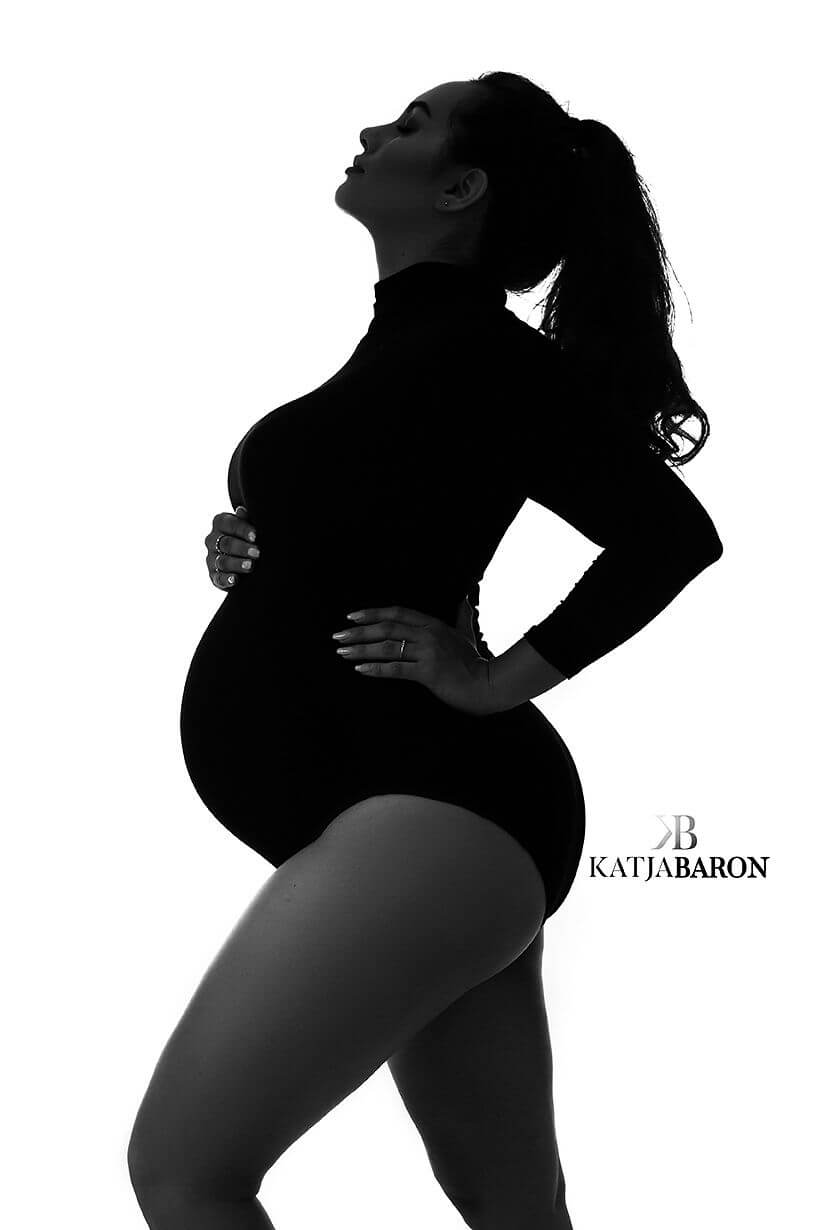 pregnant woman poses in a studio wearing a tight black bodysuit with turtleneck and long sleeves. She has a ponytail and has her face up.