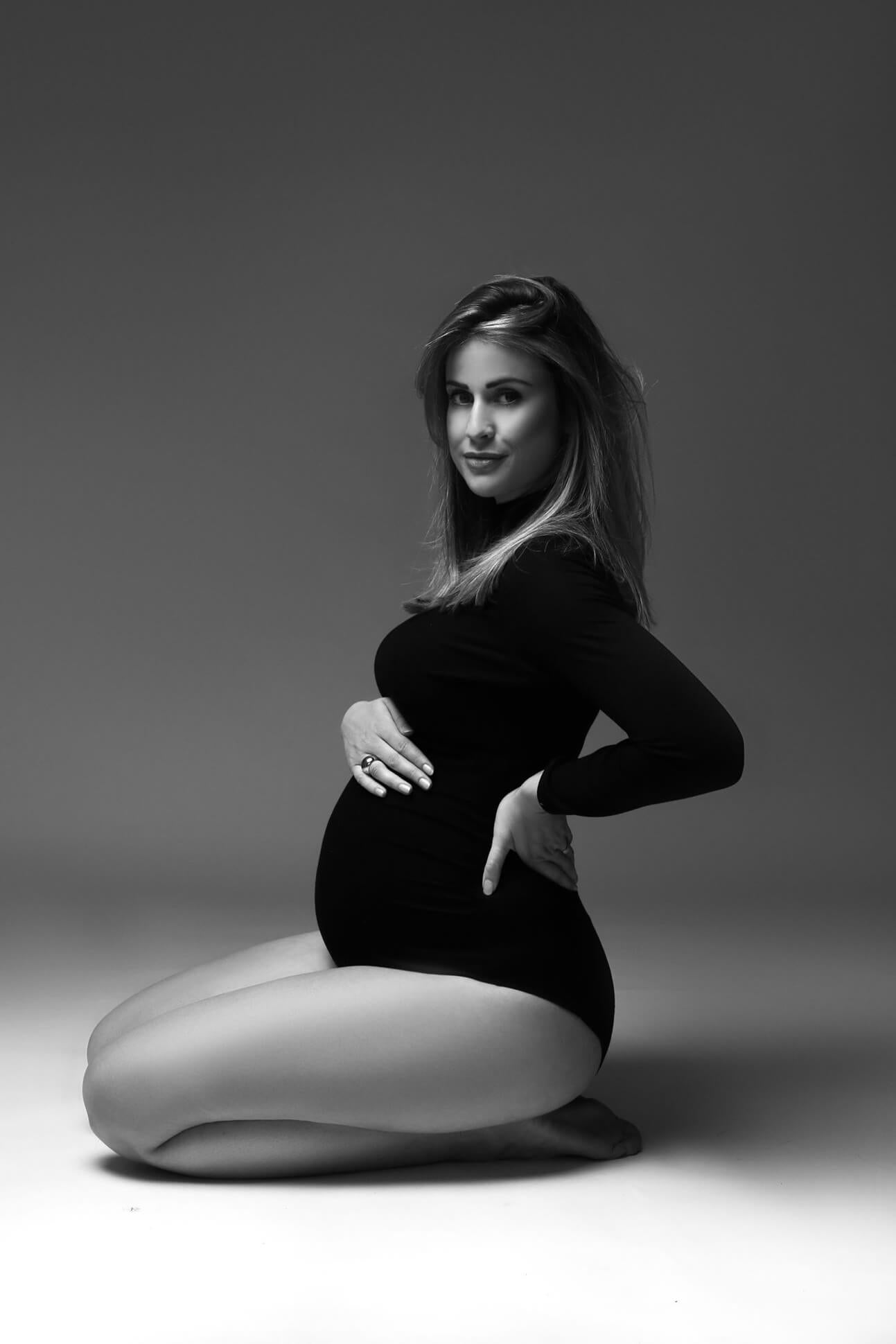 A pregnant model is wearing a black soft bodysuit. The bodysuit has a turtleneck and long sleeves. She is sitting on her knees