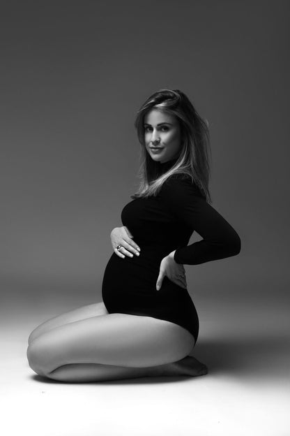 A pregnant model is wearing a black soft bodysuit. The bodysuit has a turtleneck and long sleeves. She is sitting on her knees