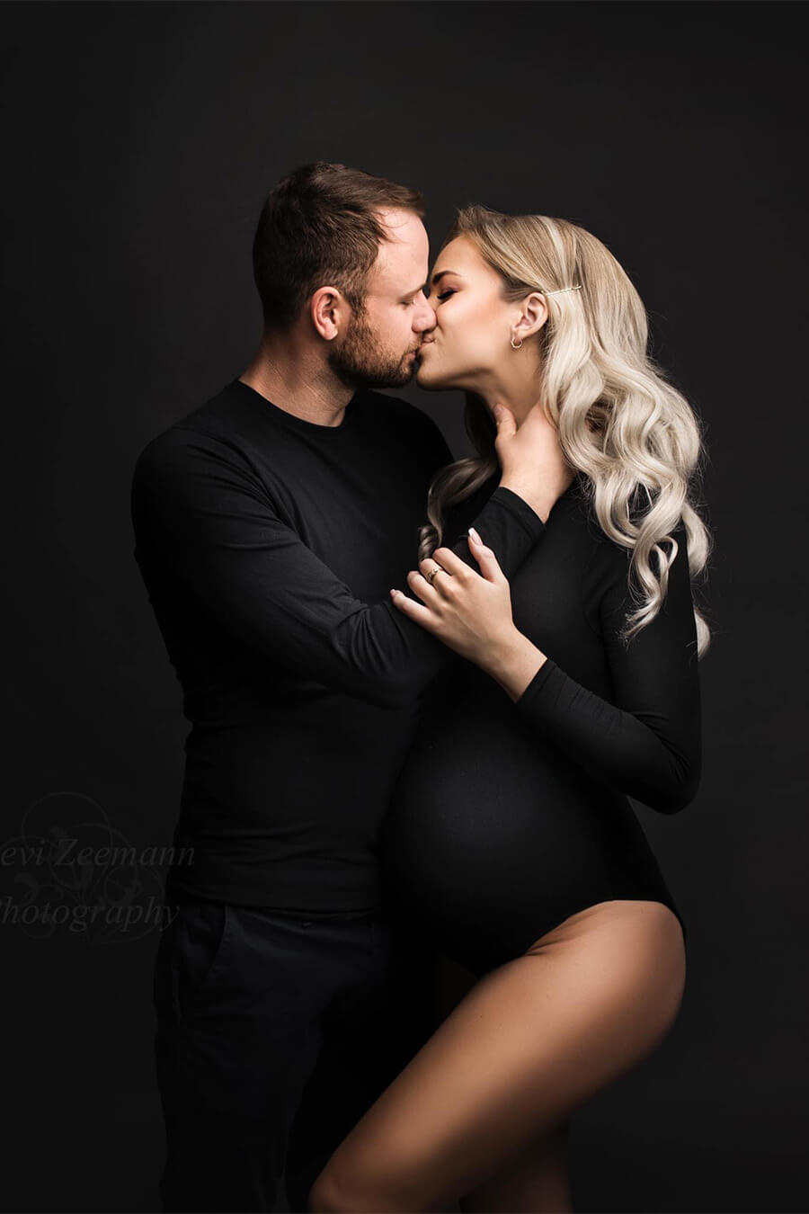couple posing in a photography studio. They are kissing each other. Both are wearing black. The male is wearing a black t-shirt with long sleeves and the woman is pregnant and wears a black bodysuit with turtleneck and long sleeves.