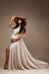 pregnant model poses in a studio with a boho styled chiffon dress in sand color. the dress has several details on the top and she also wears a brown hat to match the bohemian style. 