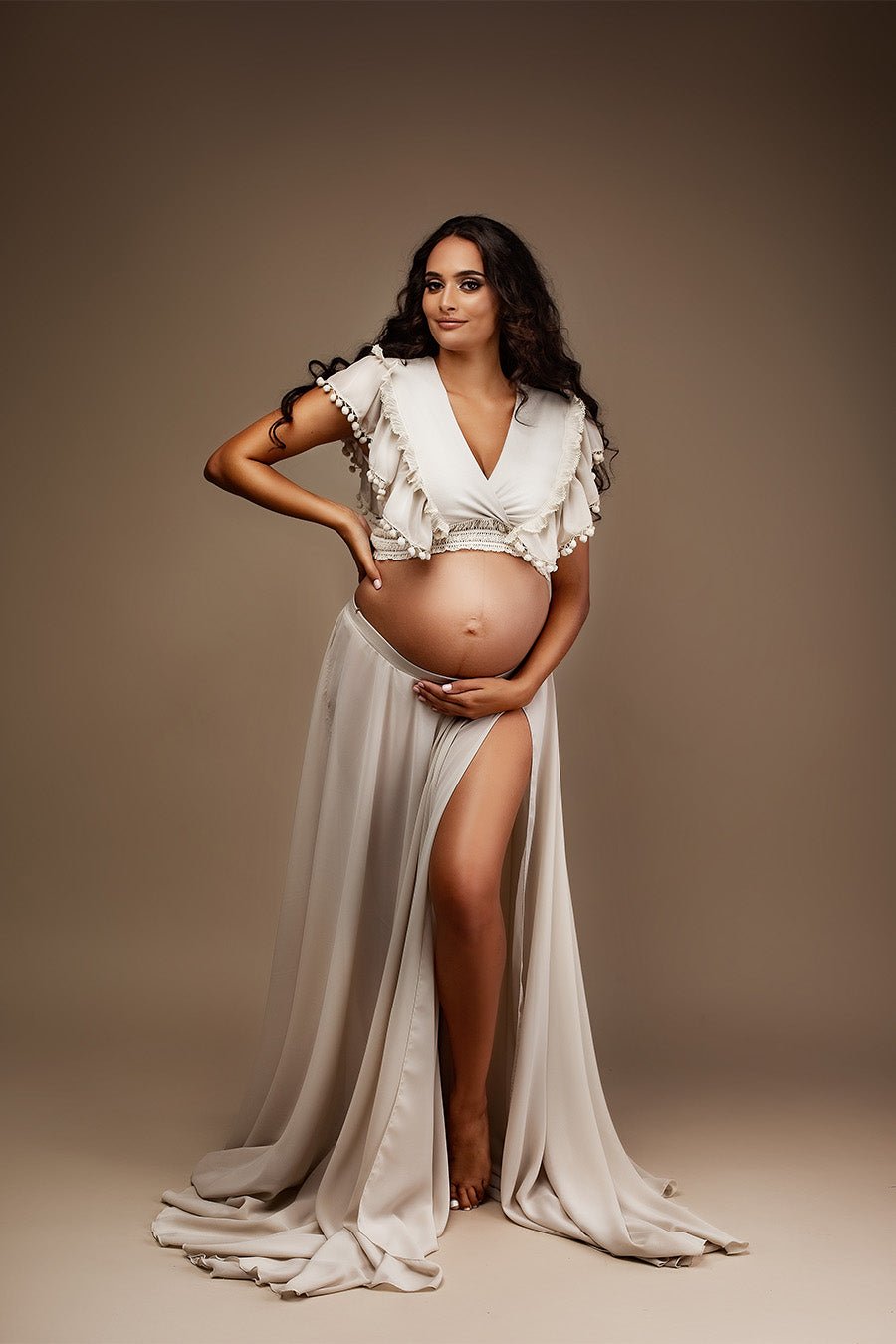 pregnant woman poses in a studio wearing a chiffon maternity set in sand color. her bely is uncovered and she is posing her hands gently in there. the top features a low v cut neckline with ruffled chiffon details. 