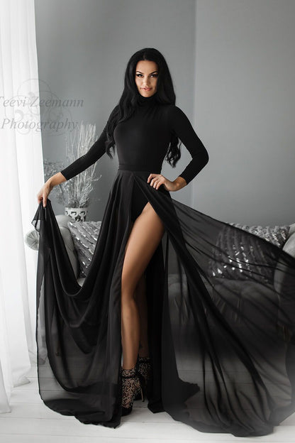 the woman is standing in a living room wearing a black bodysuit with long sleeves and a high turtleneck. The skirt is tight around her middle and is made out of a chiffon black fabric and has a split at the front