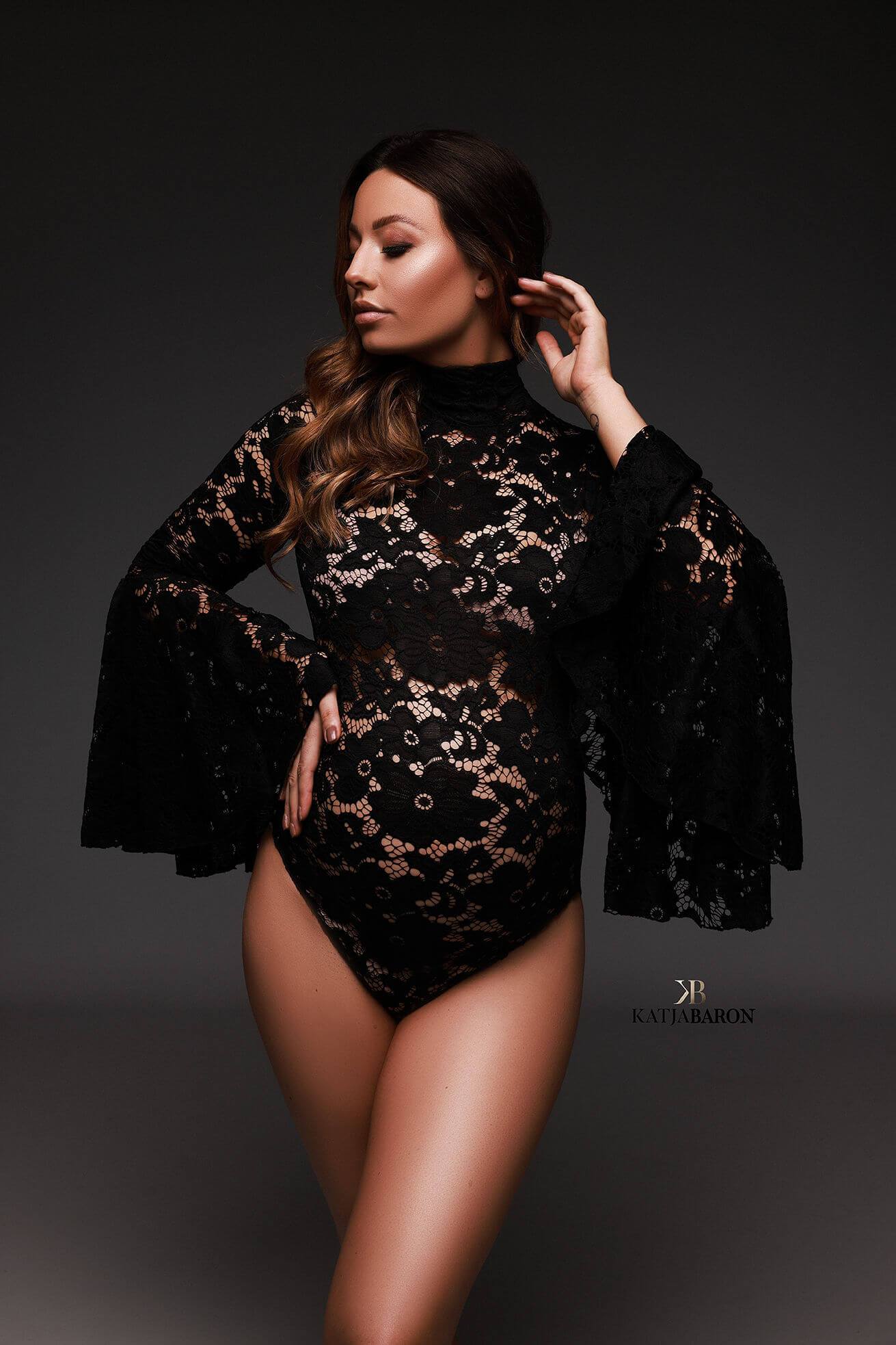 A pregnant model with brown hair is posing in a photo studio. She is wearing a black lace bodysuit. The bodysuit has a high turtle neck and wide sleeves. 