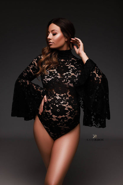 A pregnant model with brown hair is posing in a photo studio. She is wearing a black lace bodysuit. The bodysuit has a high turtle neck and wide sleeves. 