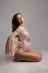 pregnant model is sitting on her knees in a studio wearing a maternity bodysuit in pink color and made off lace. the bell style sleeves are long and the top features a turtleneck. 