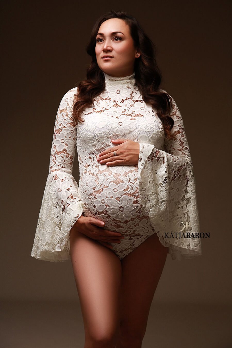A pregnant woman is standing in the studio wearing a white bodysuit.  The bodysuit is made from lace
