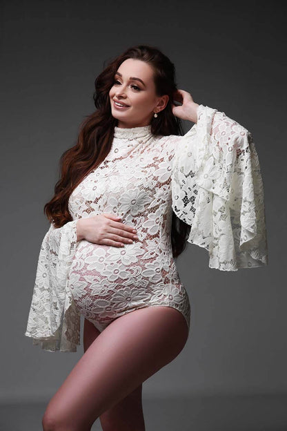 A pregnant woman with long ginger hair is posing in a photo studio. she is wearing a white bodysuit. The bodysuit has lace and a broderie flowers.