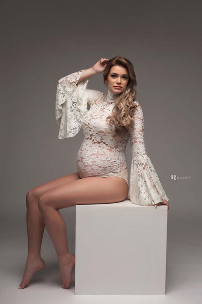 A pregnant woman is sitting on a white block in a photography studio. she is wearing a white embroidered bodysuit. She is posing with one hand in her hair and the other hand is leaning on the edge of the block.