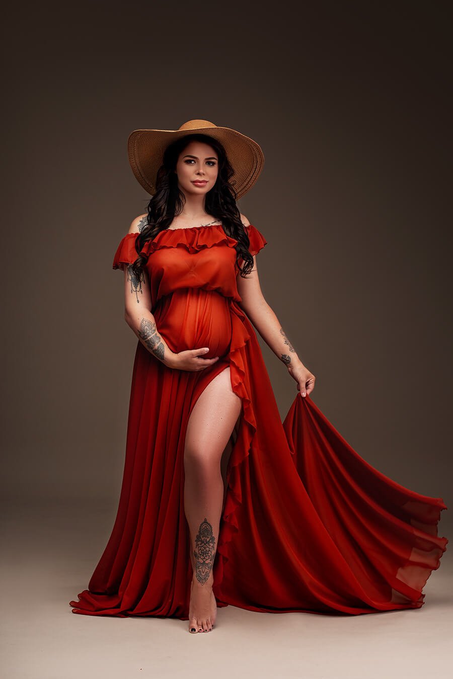 Brunette pregnant model poses for a maternity photosession in a studio. The model wears a Rust Orange colored dress and a hat to match the boho style. The dress can be completely seen in the photo - it is made of chiffon and features an off the shoulder neckline with ruffle details. The ruffle details can also been seen in the split from the circle skirt. The model has her eyes opened, one hand holding her bump and the other hand holding her skirt.