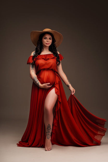 Brunette pregnant model poses for a maternity photosession in a studio. The model wears a Rust Orange colored dress and a hat to match the boho style. The dress can be completely seen in the photo - it is made of chiffon and features an off the shoulder neckline with ruffle details. The ruffle details can also been seen in the split from the circle skirt. The model has her eyes opened, one hand holding her bump and the other hand holding her skirt.