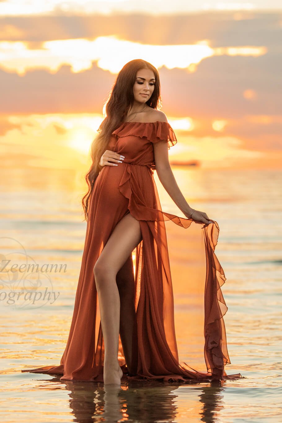 Brunette pregnant model poses for a maternity photosession outside on the beach. She stands with her feet in the water and wears a Mii-Estilo Florian Dress in Rust Orange color. The dress can be completely seen in the photo - it is made of chiffon and features an off the shoulder neckline with ruffle details. The ruffle details can also been seen in the split from the circle skirt. The model has her eyes closed, one hand holding her bump and the other hand holding her skirt. 
