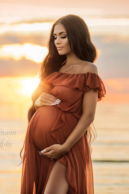 Brunette pregnant model poses for a maternity photo session at the beach. She wears a Mii-Estilo Florian Dress in Rust Orange color. The dress can be partially seen in the photo - it is made of chiffon and features an off the shoulder neckline with ruffle details. The ruffle details can also been seen in the split from the circle skirt. The model has her eyes closed and holds her bump with both hands.