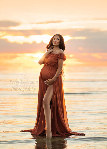 Brunette pregnant model poses for a maternity photosession outside on the beach. She stands with her feet in the water and wears a Mii-Estilo Florian Dress in Rust Orange color. The dress can be completely seen in the photo - it is made of chiffon and features an off the shoulder neckline with ruffle details. The ruffle details can also been seen in the split from the circle skirt. The model looks to the camera and has one hand holding her bump and the other hand laying on her neck.