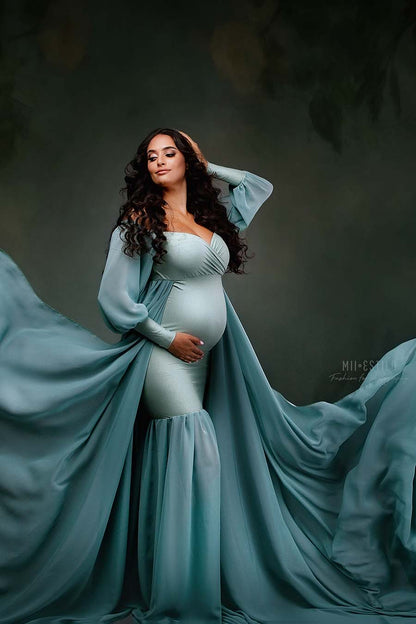pregnant model poses in a studio wearing a long dress made of jersey and chiffon in azur color. she touches her hair and bump while posing. the dress features a chiffon train with lots of fabric to create movement. the top has an adjustable sweetheart neckline, off shoulder and chiffon bishop sleeves. 