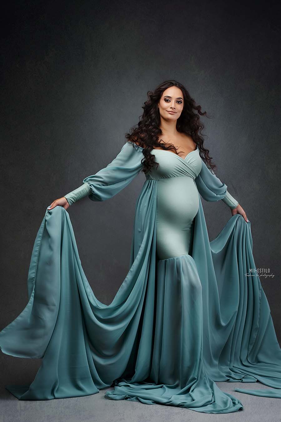 model poses during a pregnancy photoshoot. she wears a dress in azur color. the dress features a chiffon train with lots of fabric to create movement. the top has an adjustable sweetheart neckline, off shoulder and chiffon bishop sleeves.