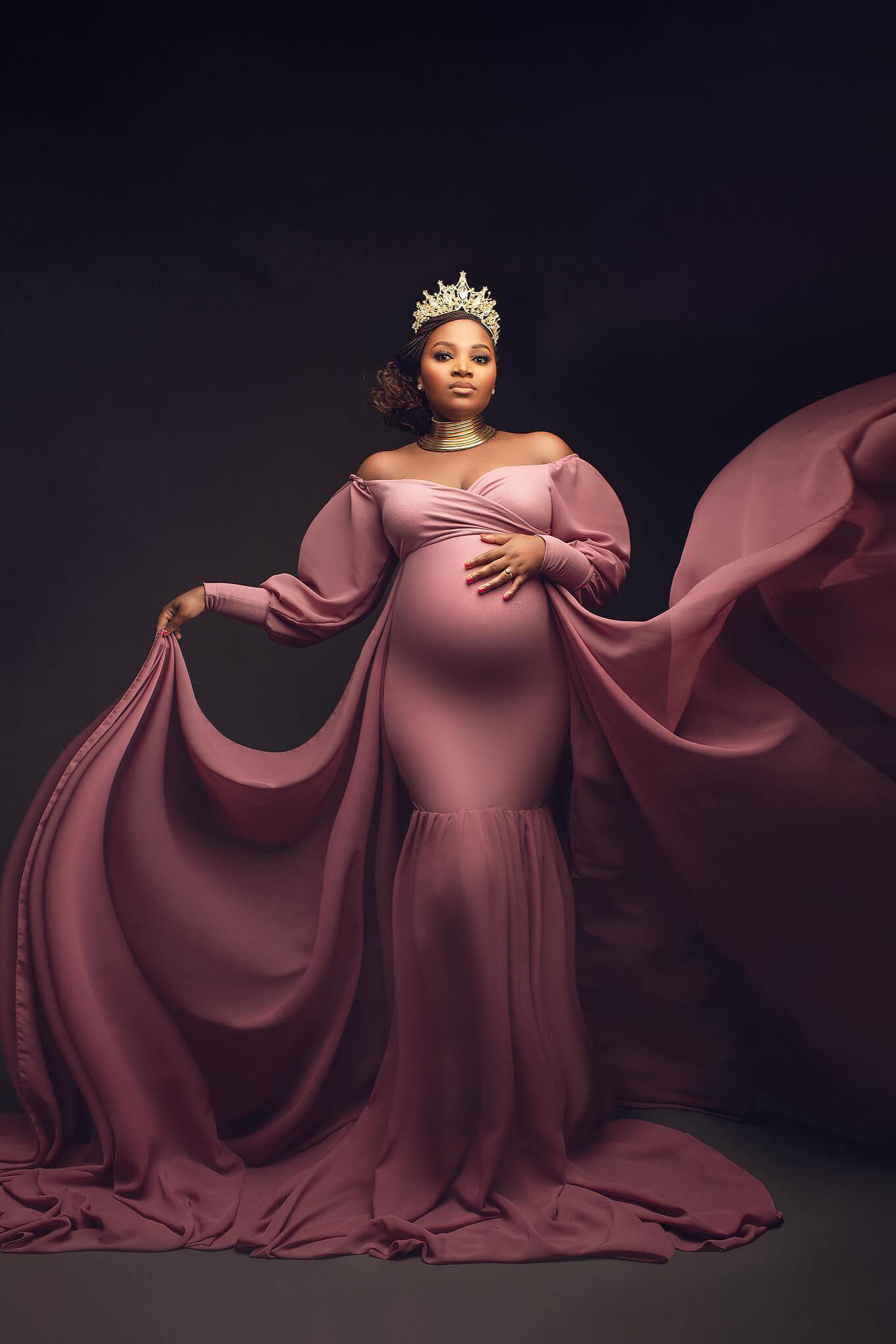 black pregnant model poses in a studio dresses with a long dress made of chiffon and jersey. she has a crown and a large necklace to match the style. the dress features an adjustable sweetheart top, off shoulder top and long chiffon bishop sleeves with jersey manchettes. the dress is tight, made of jersey and the skirt is made of chiffon. there is a chiffon train attached under the chest.