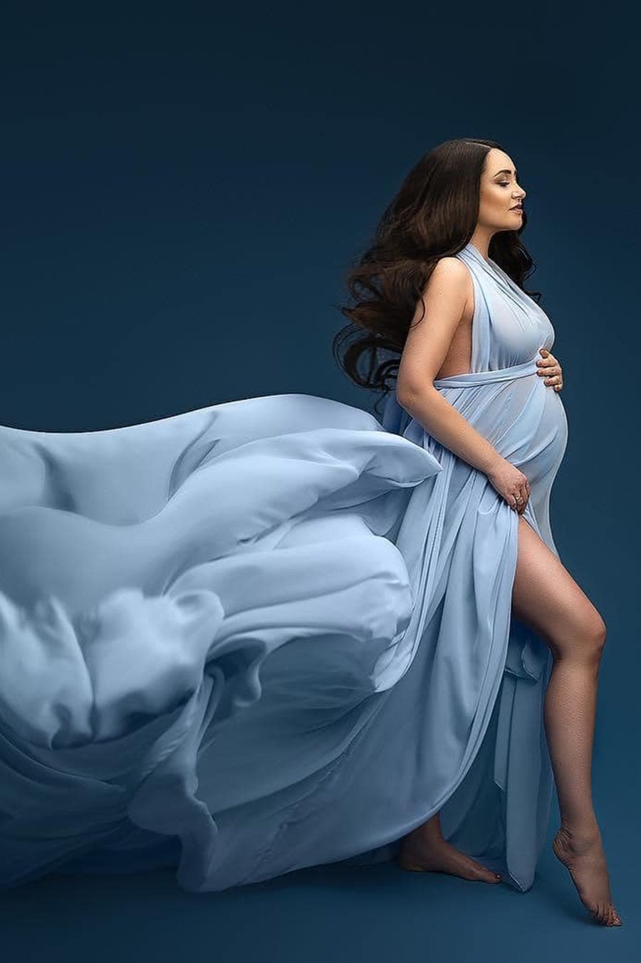 A pregnant woman is wearing a long blue dress. The dress is made from chiffon with a lot of fabric. The fabric is used to created waves behind her