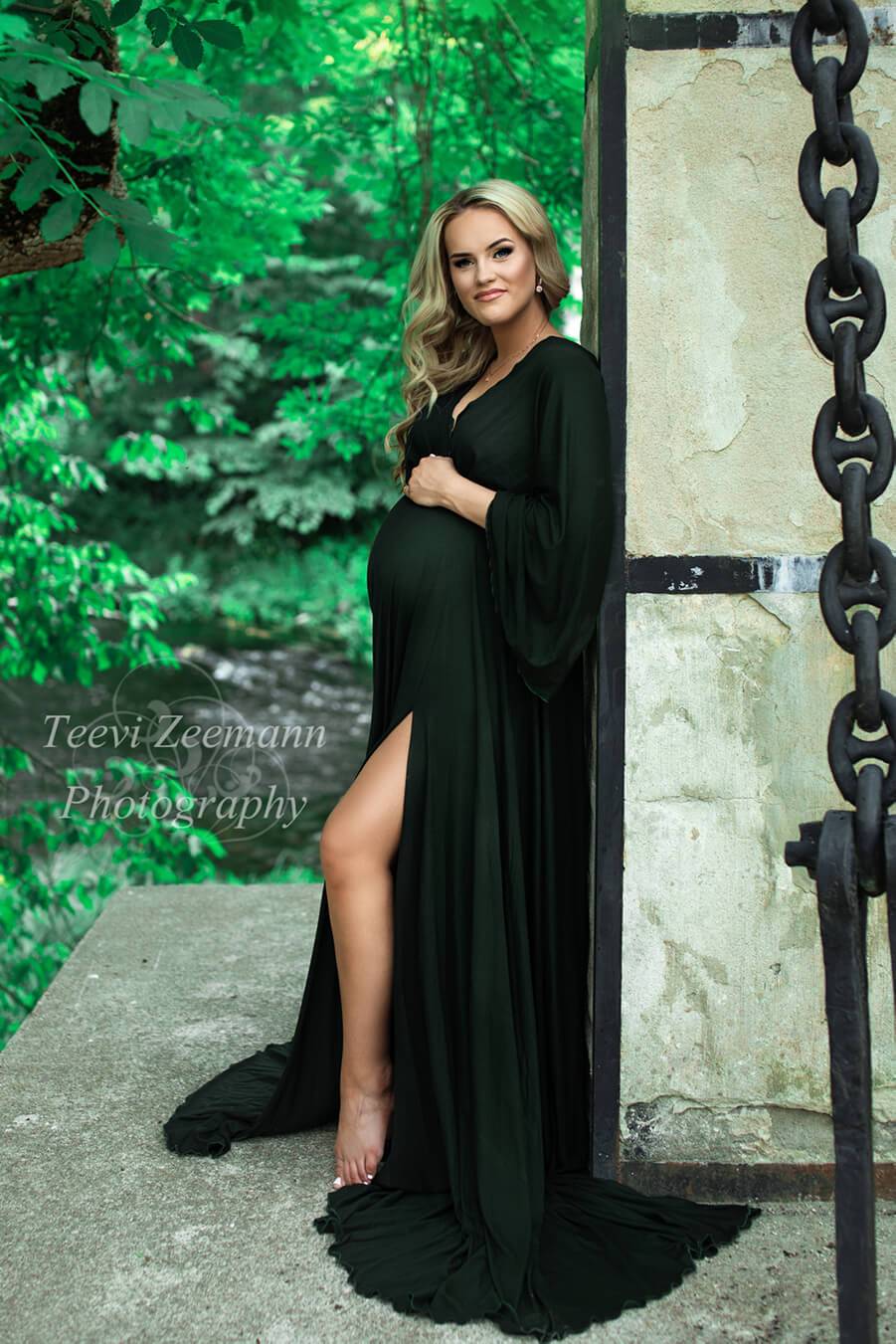 This pregnant model is leaning against a wall in a garden. She is wearing a dark green maternity dress with a split by the leg. She has her hands on her belly