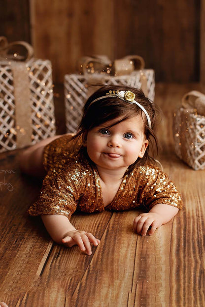 A baby girl is laying on the ground. There are little golden present in the background. The babygirl is wearing a gold romper with sequins. The girl is looking to the camera and has a little smile.