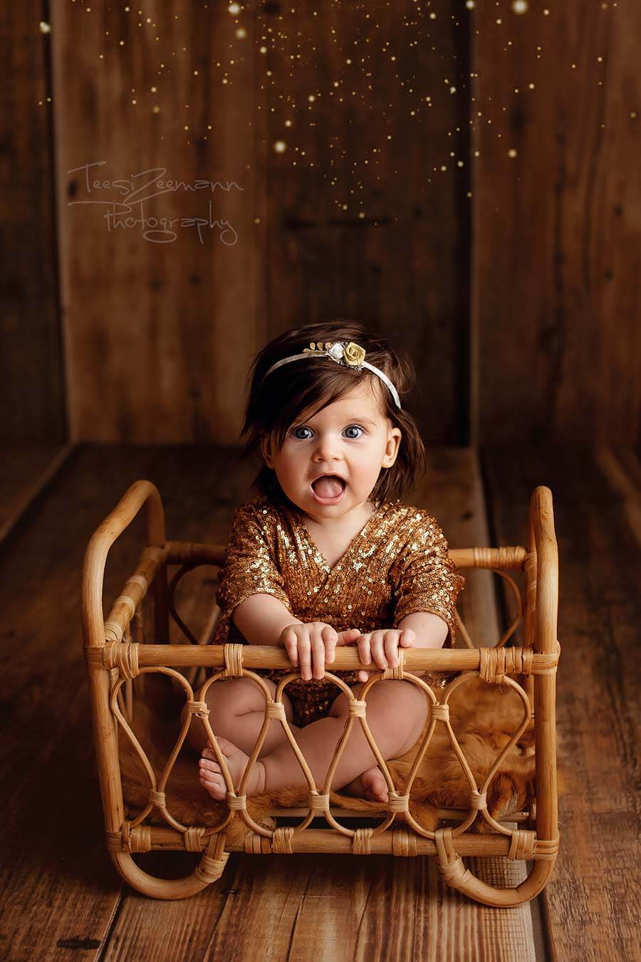 A baby girl is sitting in a small bed. She is wearing a golden romper. the romper is made out of gold sequins for that sparkly look. She has a little hairband with a small rose on it in her hair. 