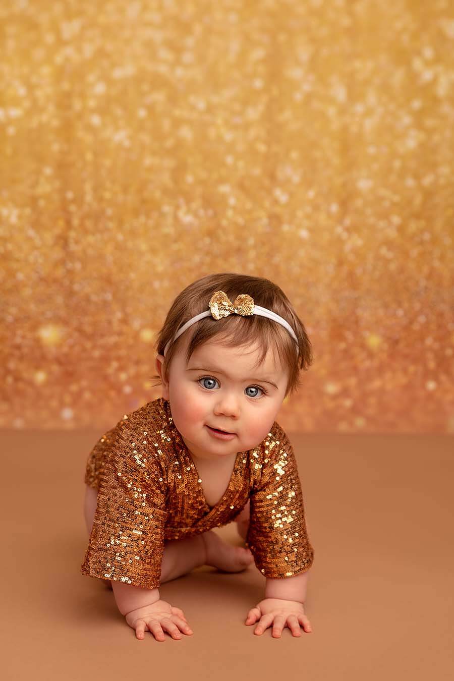 A baby girl is crawling on the floor. She is wearing a cute baby romper with gold sequins on it. she has a matching gold bow in her hair. The background has gold glitters.\