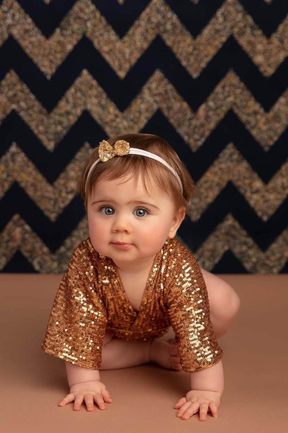 A babygirl is Crawling on the floor. She has a little bow in her hair . She is wearing a golden romper with sequins on it for a sparkly look. 