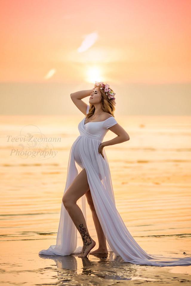 blond pregnant model poses outside by the water wearing a white dress with short sleeves and off the shoulder neckline. the skirt is made of chiffon. 