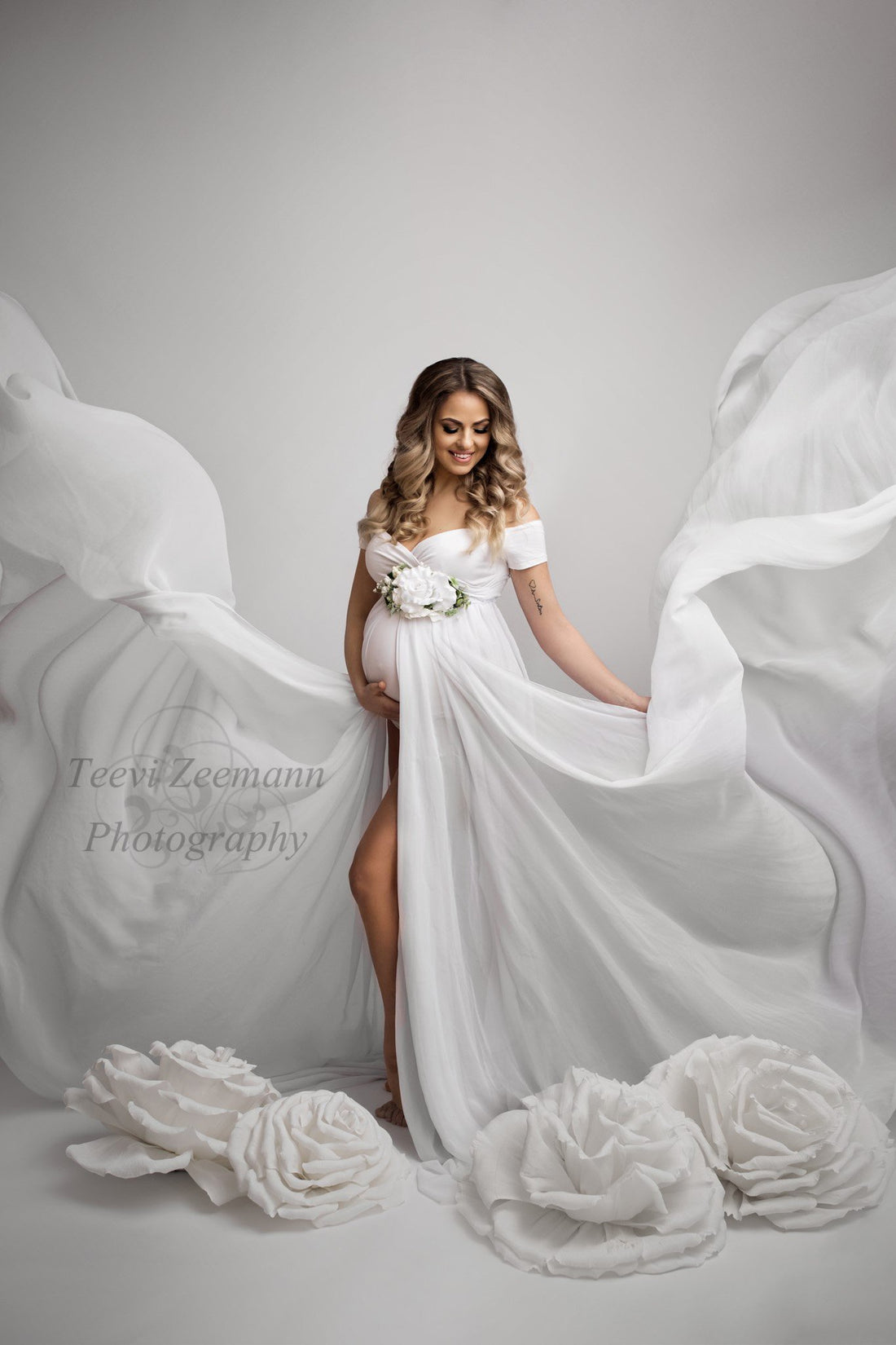 blond pregnant model poses in a studio wearing a materntiy dress with long chiffon trains. the trains do give an incredible butterfly effect on the photo. the model has a white flower on the waist under the top. 
