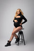 blond model poses sitting on a high chair and wearing a black jersey bodysuit with long sleeves, keyhole neck and a turtleneck neckline. 
